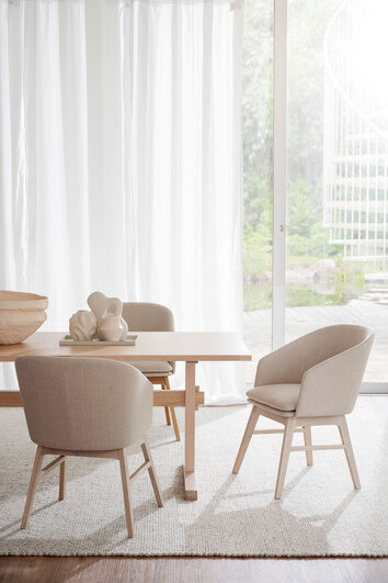 Windhamn Karmstol i vitpigmenterad ek, klädd i beige tyg. Karmstolen står vid ett rektangulärt matbord i ljust trä.