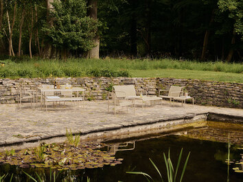 Loungeserien Dapple i färgen Cashmere som står på en stenig uteplats bredvid en damm