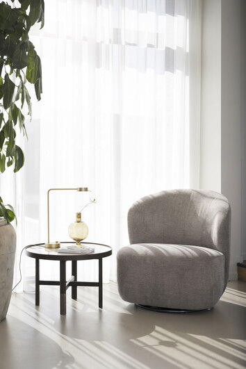 Roselawn Fåtölj vid fönster med skira gardiner bredvid ett runt sidobord med bordslampa och vas