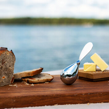 Tilt Kniv Stål på skärbräda med bröd och ost. I bakgrunden syns hav och skog i horisonten.