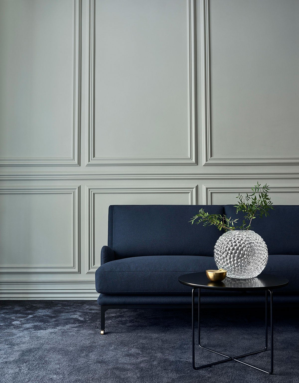 Miljöbild på soffan Mr. Jones klädd i mörkblå tyg Rico 140. Ramen är av svart stål, med hjul i mässing. 