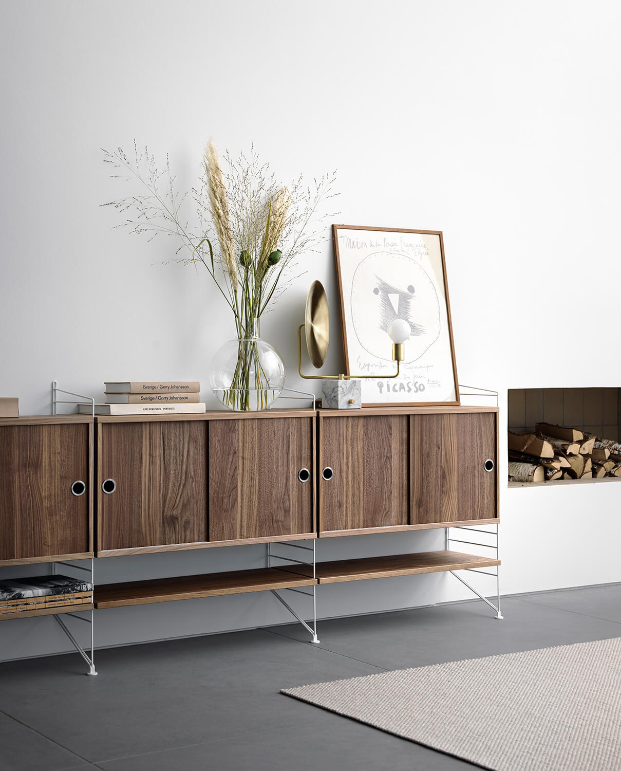 Stringhylla bestående av skåp med skjutdörrar och hyllplan i valnöt ståendes i vardagsrum med tillhörande inredning.
