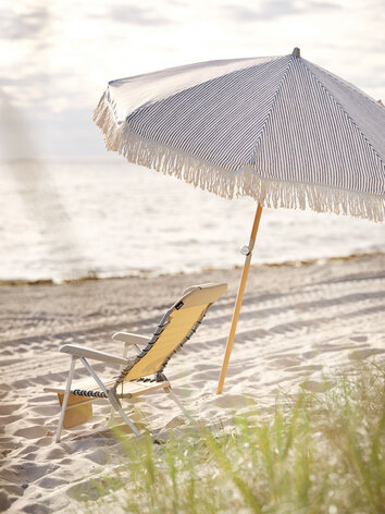 Gatsby Parasoll Blå / Vit på stranden bredvid en gul solstol