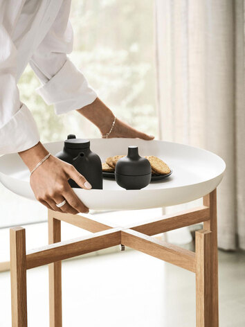 Tablo Bord i hög modell. En kvinna lyfter bordsskivan från stativet. Bordsskivan är dekorerad med kex och en tekanna.