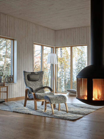 Easy Fåtölj med fotpall i fårskinnet Graphite, bredvid en tänd brasa