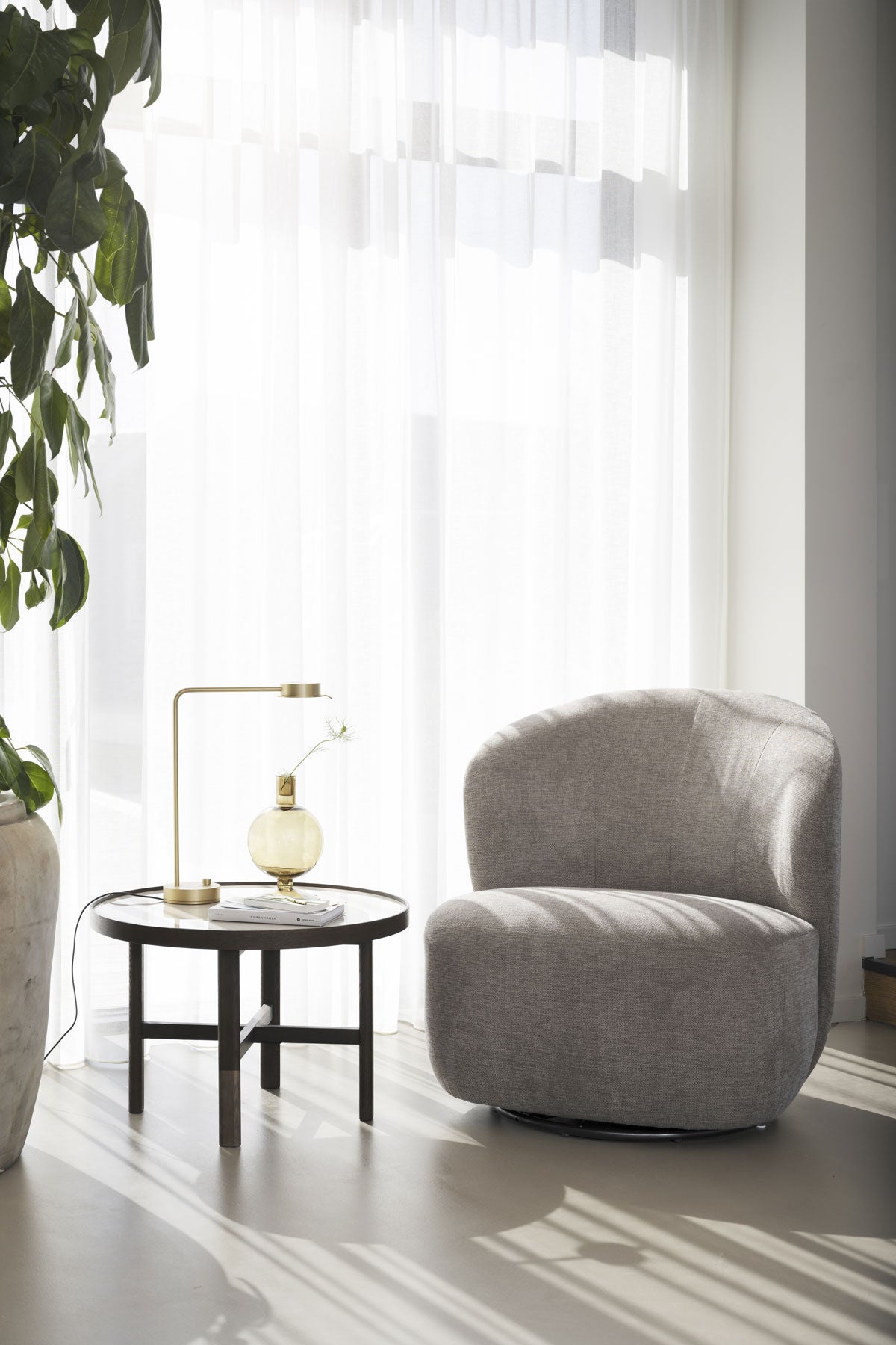Roselawn Fåtölj vid fönster med skira gardiner bredvid ett runt sidobord med bordslampa och vas