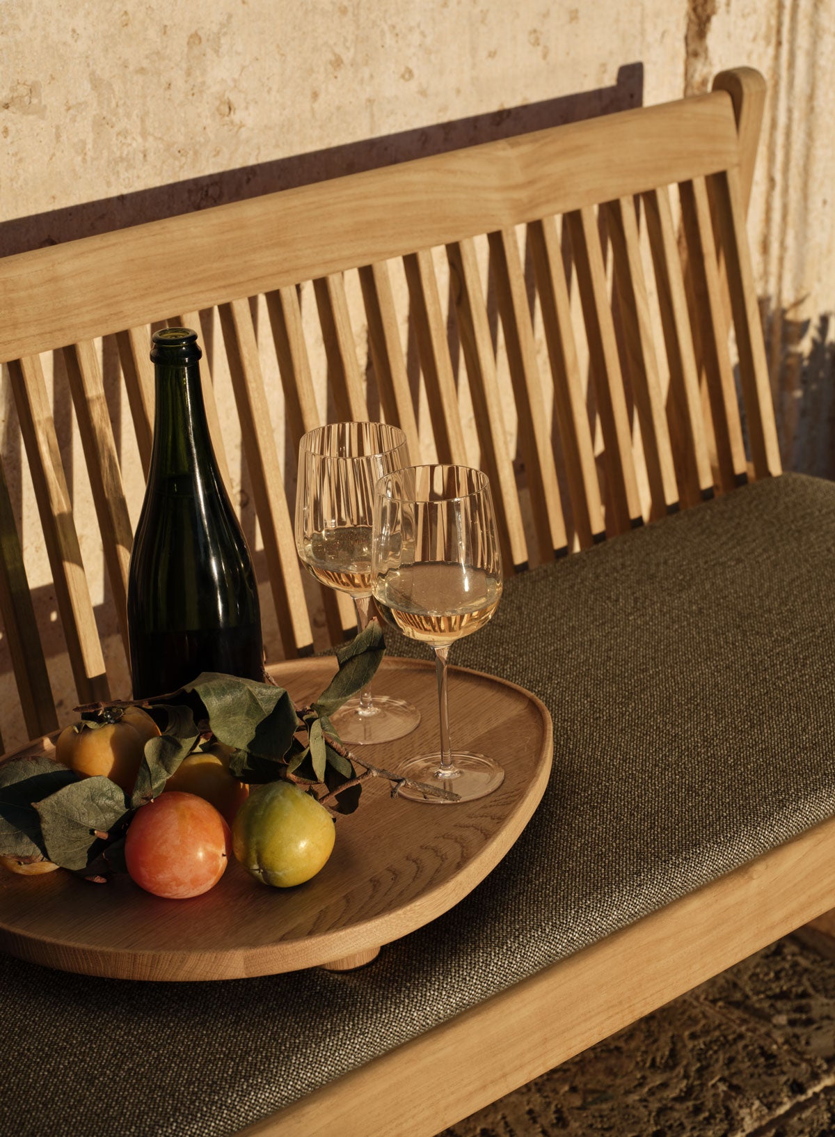 Patio trädgårdsbänk i solnedgång med en bricka med citrusfrukter, en flaska vin och två halvfyllda vinglas.