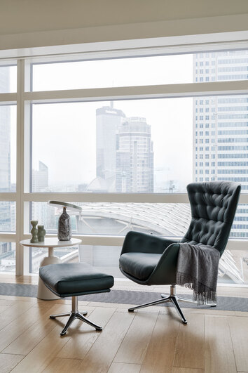 Sini Fåtölj och fotpall i lädret Savoy Deep Forest Green med ben i polerad aluminium framför ett fönster som vetter ut mot skyskrapor