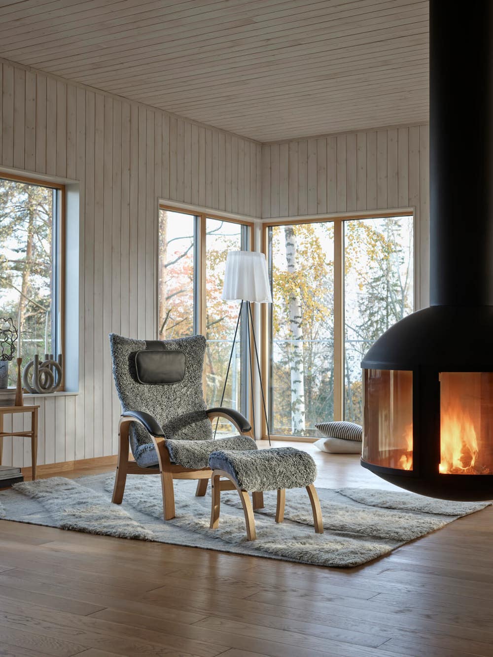 Easy-fatolj-Conform_2023-miljo01.jpg Easy Fåtölj med fotpall i fårskinnet Graphite, bredvid en tänd brasa