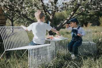Barr Loungestol och Barr Loungestol Mini i sommardröm. Loungegruppen står i en blommande äppleträdgård och barn som fikar på jordgubbstårta.